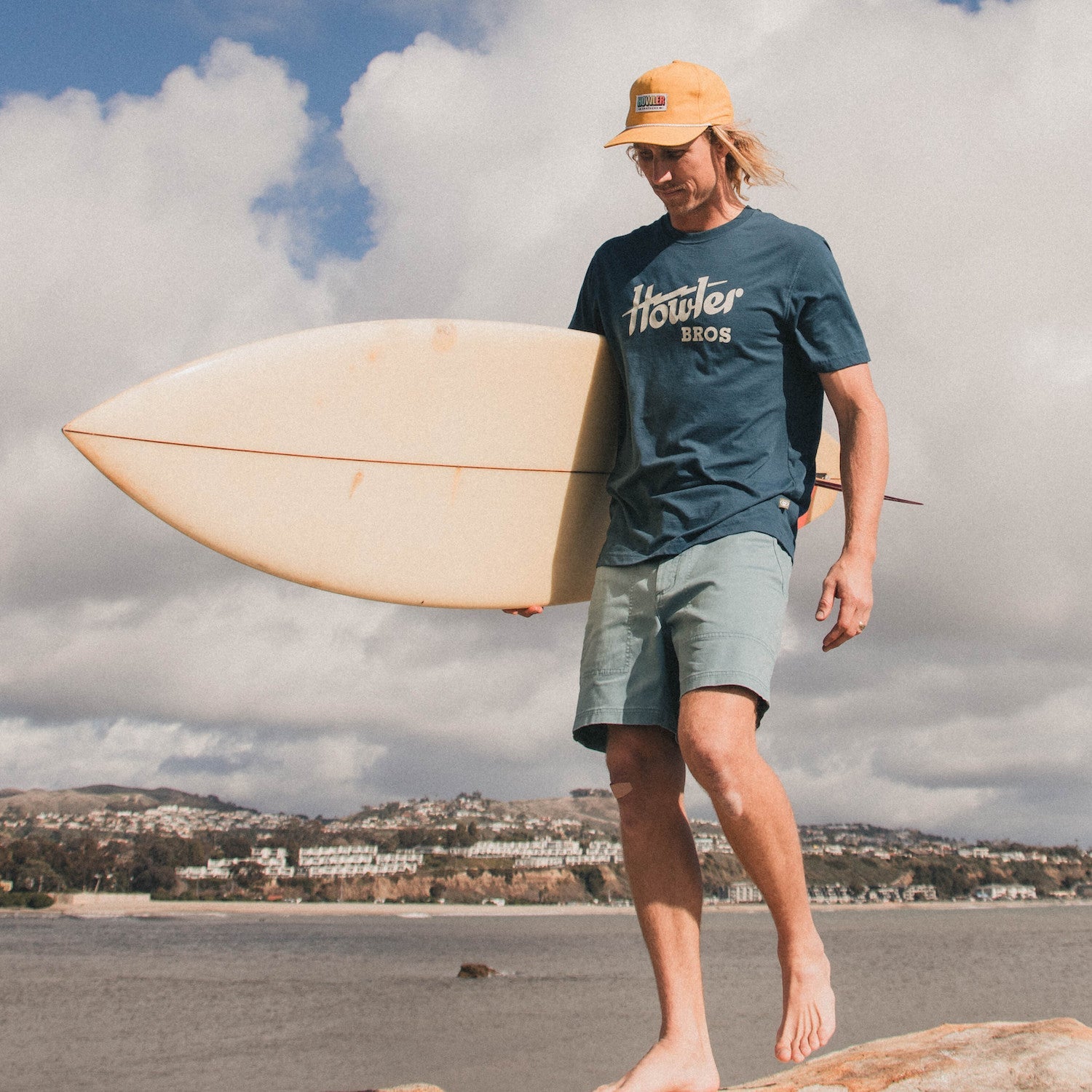 A man wearing a yellow cap, classic Howler designs blue cotton-poly blend t-shirt, and light green shorts walks barefoot on rocks by the water, carrying a surfboard with a coastal town and cloudy sky in the background.