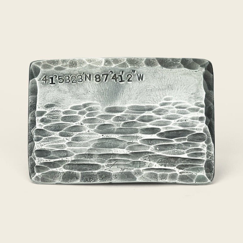 A rectangular metallic custom coordinates buckle with a textured, hammered surface featuring engraved 41°5323N 87°412W in the top left corner, crafted by Cityboy Forge, set against a neutral background.