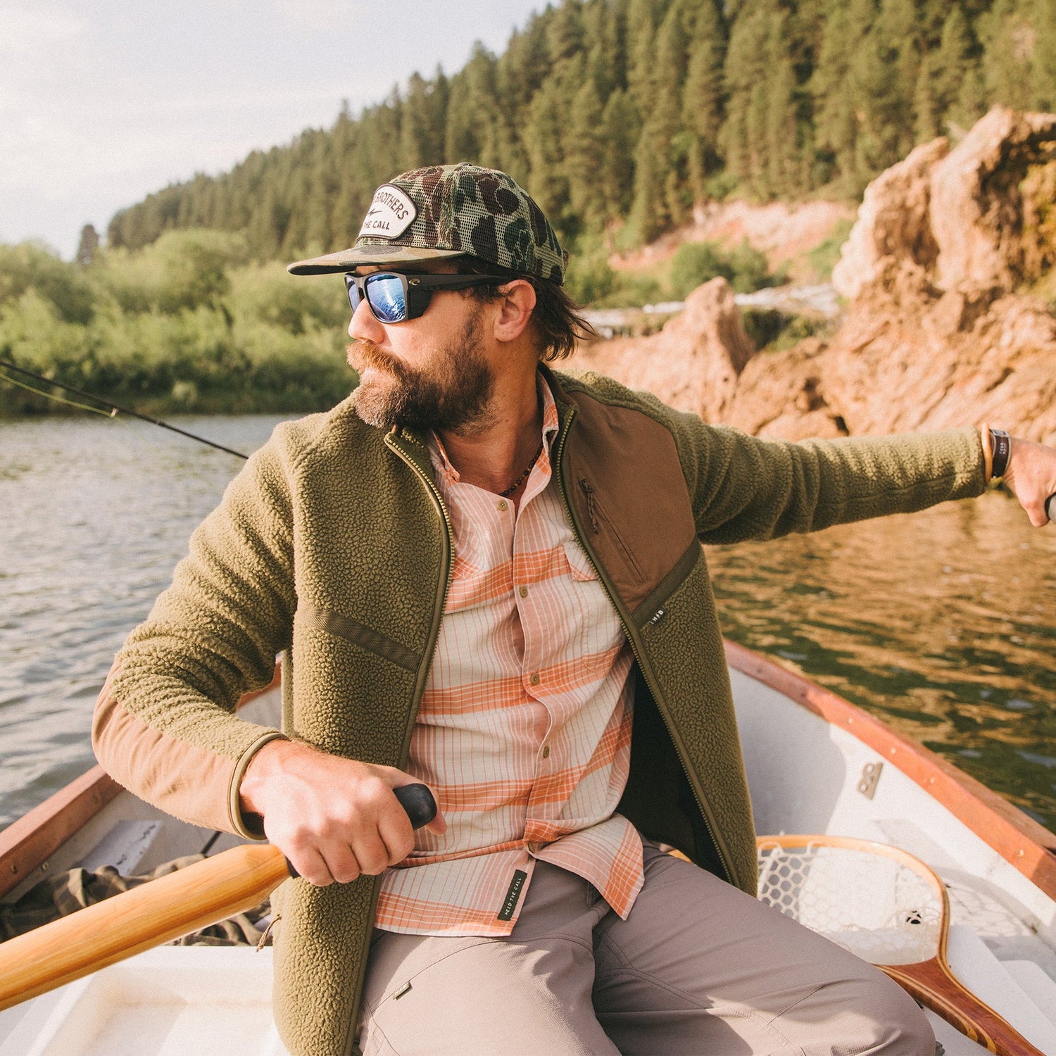 A man wearing sunglasses, a cap, and a fleece jacket rows a small boat on a lake surrounded by trees and rocky hills, looking to the side.
