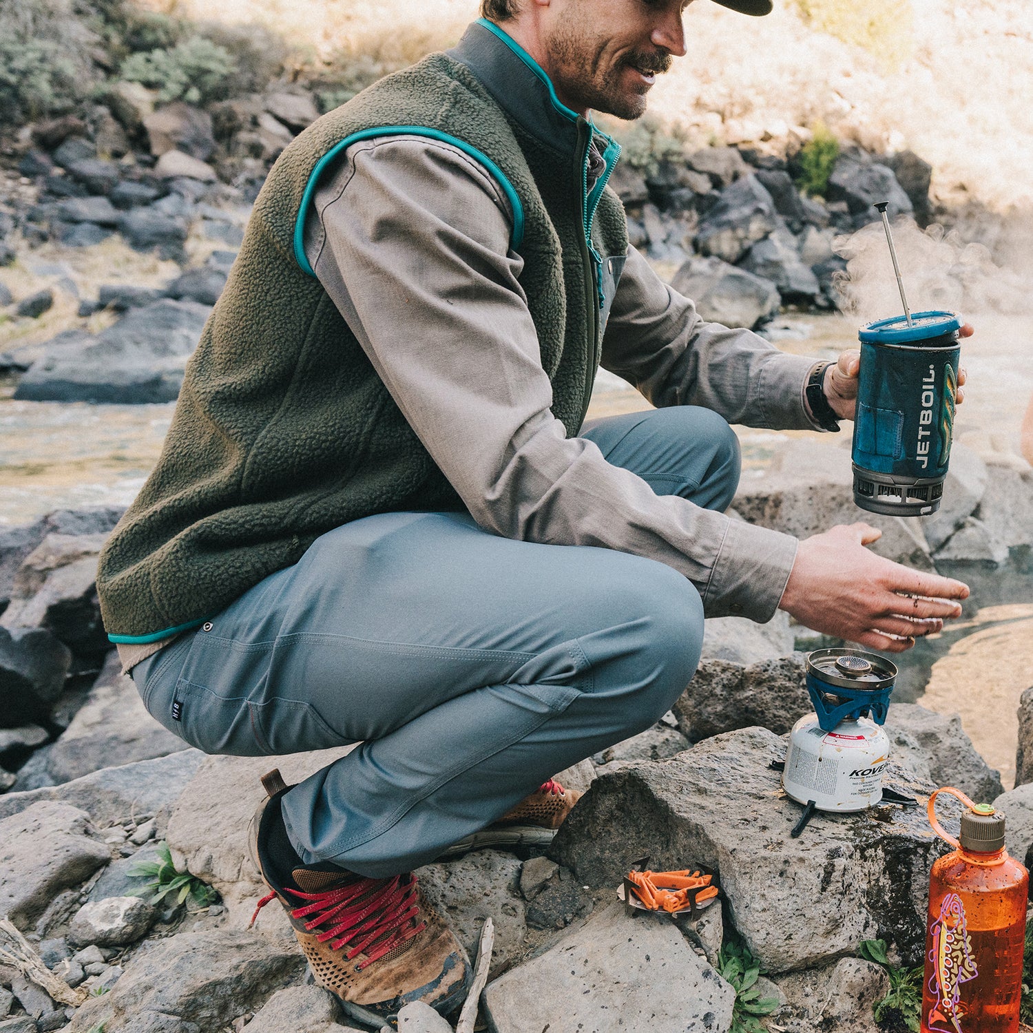 A person in outdoor gear and tech fabric pants kneels on rocks by a river, warming their hands over a portable camping stove with a blue Jetboil cooking system. An orange fuel canister and multitool are nearby.