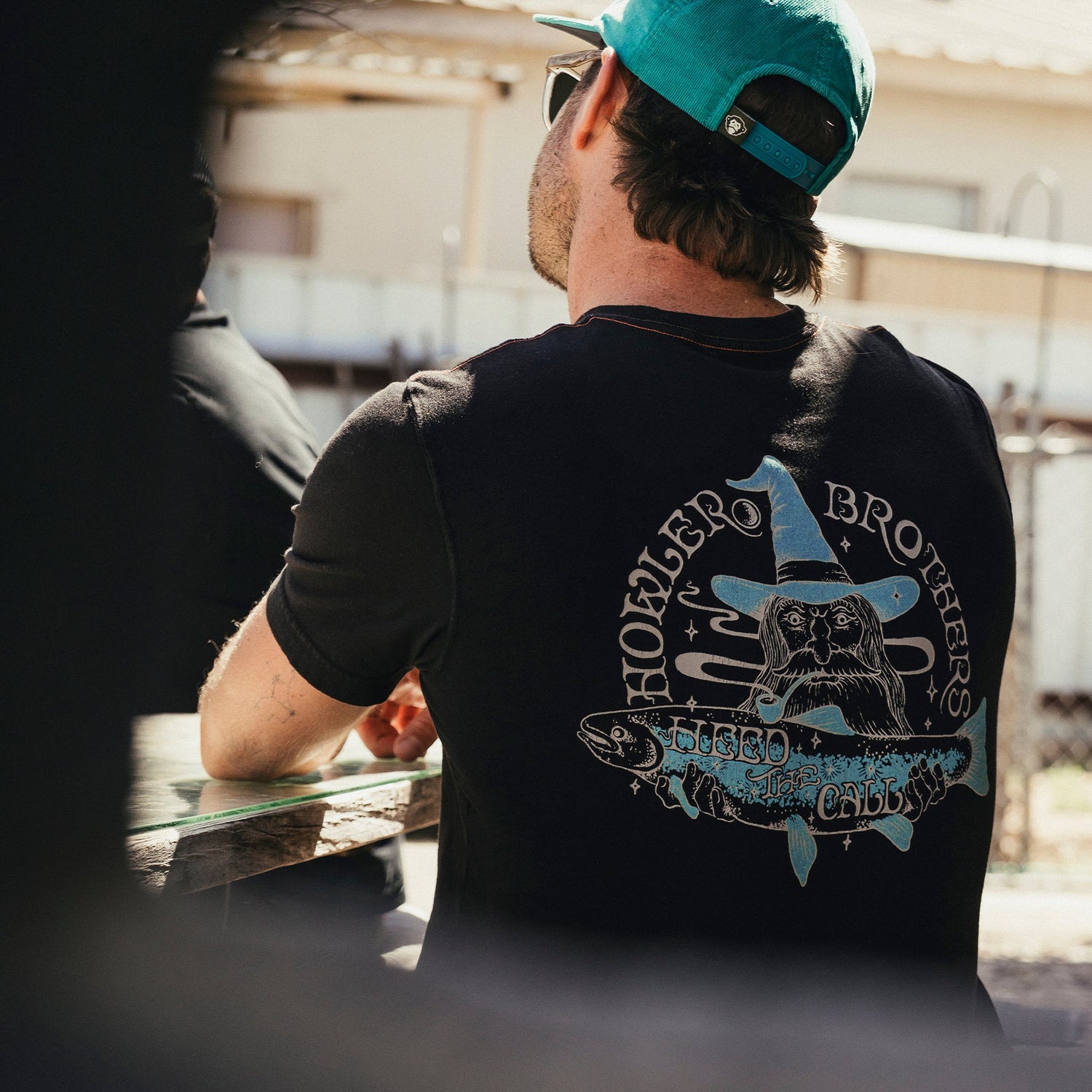 A man wearing a turquoise cap and a black cotton t-shirt with a graphic wizard hat, wolf, and the text “Howler Brothers” sits outdoors at a picnic table, facing away from the camera, capturing a subtle vintage look.