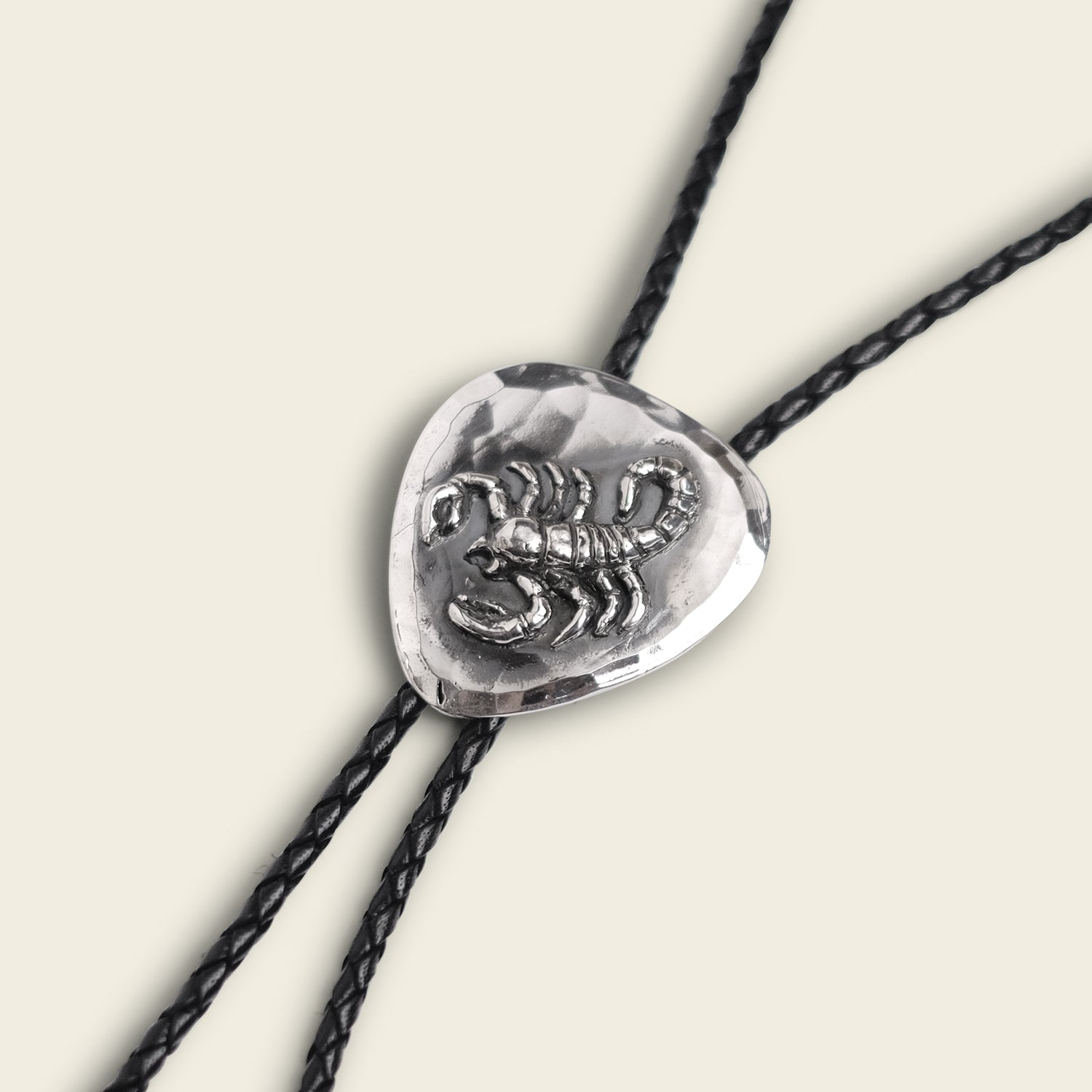 A silver El Alacrán Bolo Tie slide with a raised scorpion design is threaded onto two black braided leather cords, set against a light background—an eye-catching handmade bolo tie by Cityboy Forge.