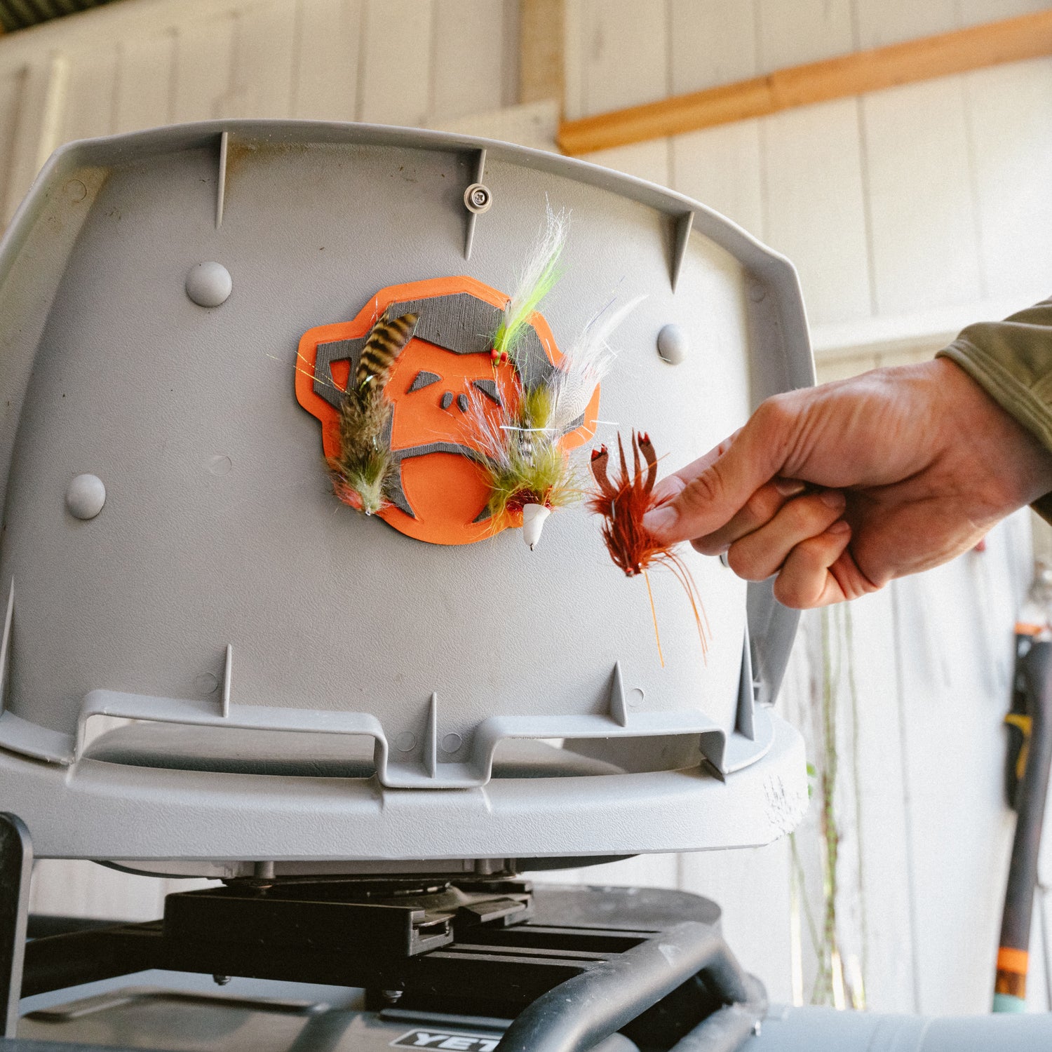 A hand places colorful fishing flies onto the El Mono Hook Pad, a high-density foam fly holder attached to a gray container inside a shed or garage.