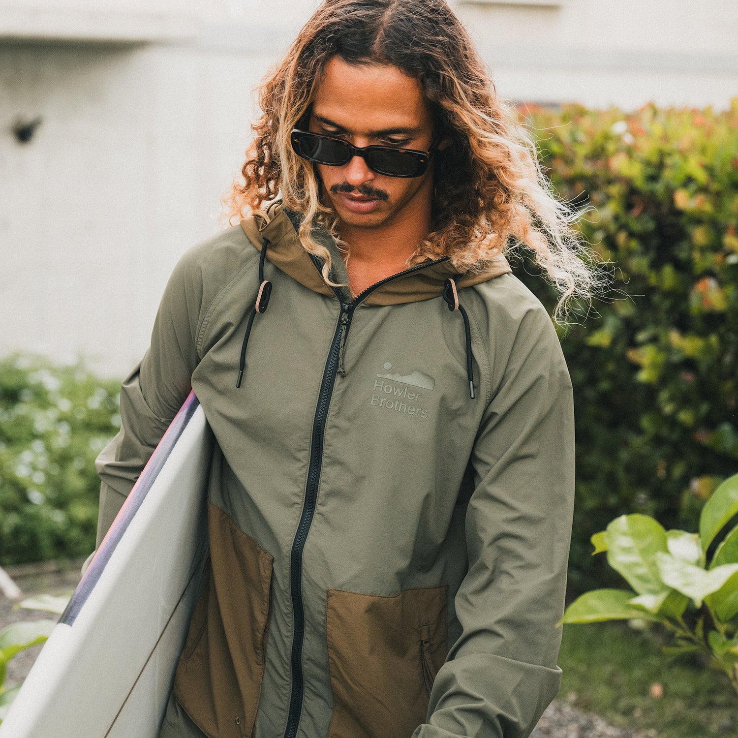 A person with long, wavy hair wearing sunglasses and a green lightweight windbreaker holds a surfboard while standing outdoors near greenery.