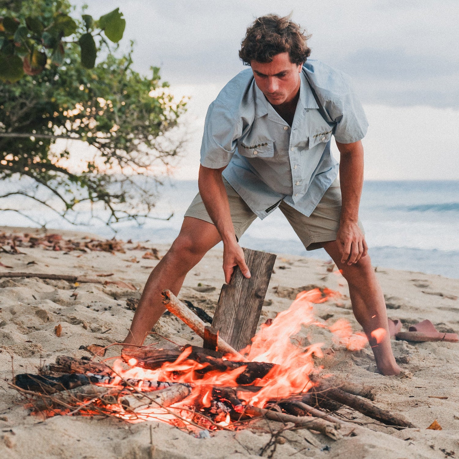 A man in a light blue shirt and cotton twill shorts adds a piece of wood to a campfire on a sandy beach, with the ocean and trees visible in the background.