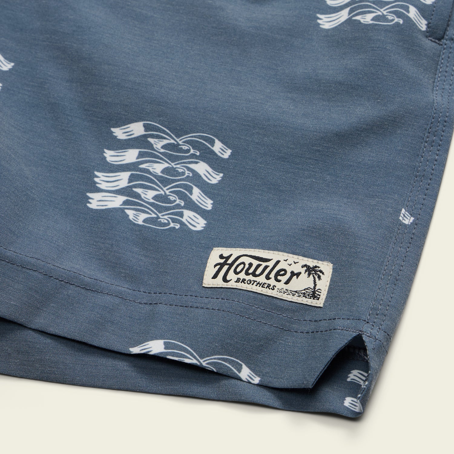 Close-up of blue swim trunks featuring white seagull patterns, an elastic waistband, and a stitched Howler Brothers label on the hem with a palm tree graphic. - View 6