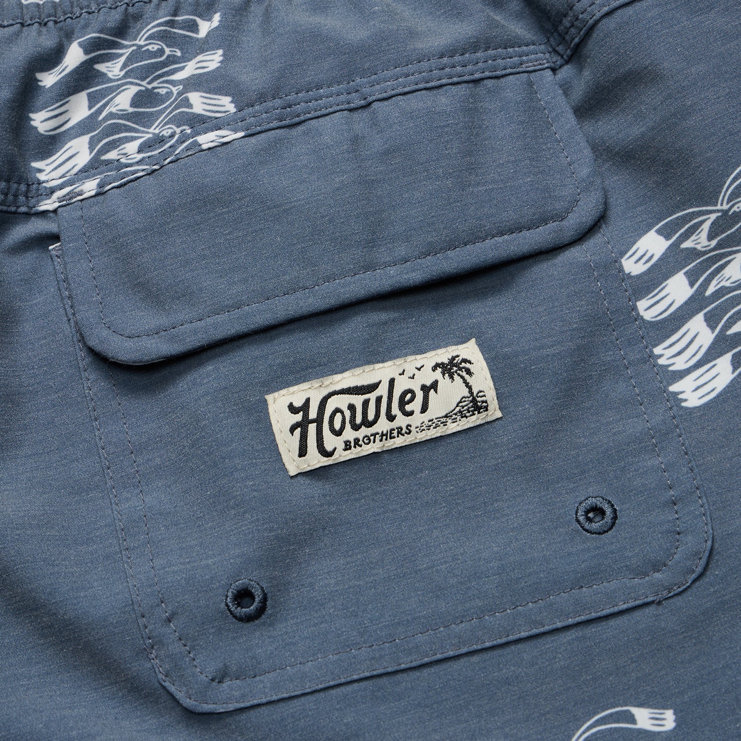 Close-up of a blue fabric pocket with a Howler Brothers label sewn on, seen on their boardshorts. The fabric features a white bird pattern and two metal eyelets at the bottom of the pocket. - View 7