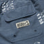Close-up of a blue fabric pocket with a Howler Brothers label sewn on, seen on their boardshorts. The fabric features a white bird pattern and two metal eyelets at the bottom of the pocket.