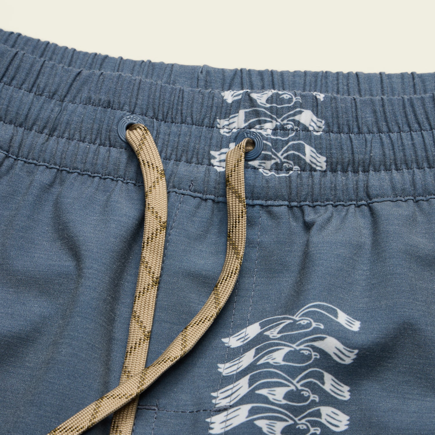 Close-up of blue boardshorts featuring a beige drawstring, an elastic waistband, and a repeating white seagull pattern along the waistband and side seam. 7” inseam offers comfort and style for any beach day. - View 5