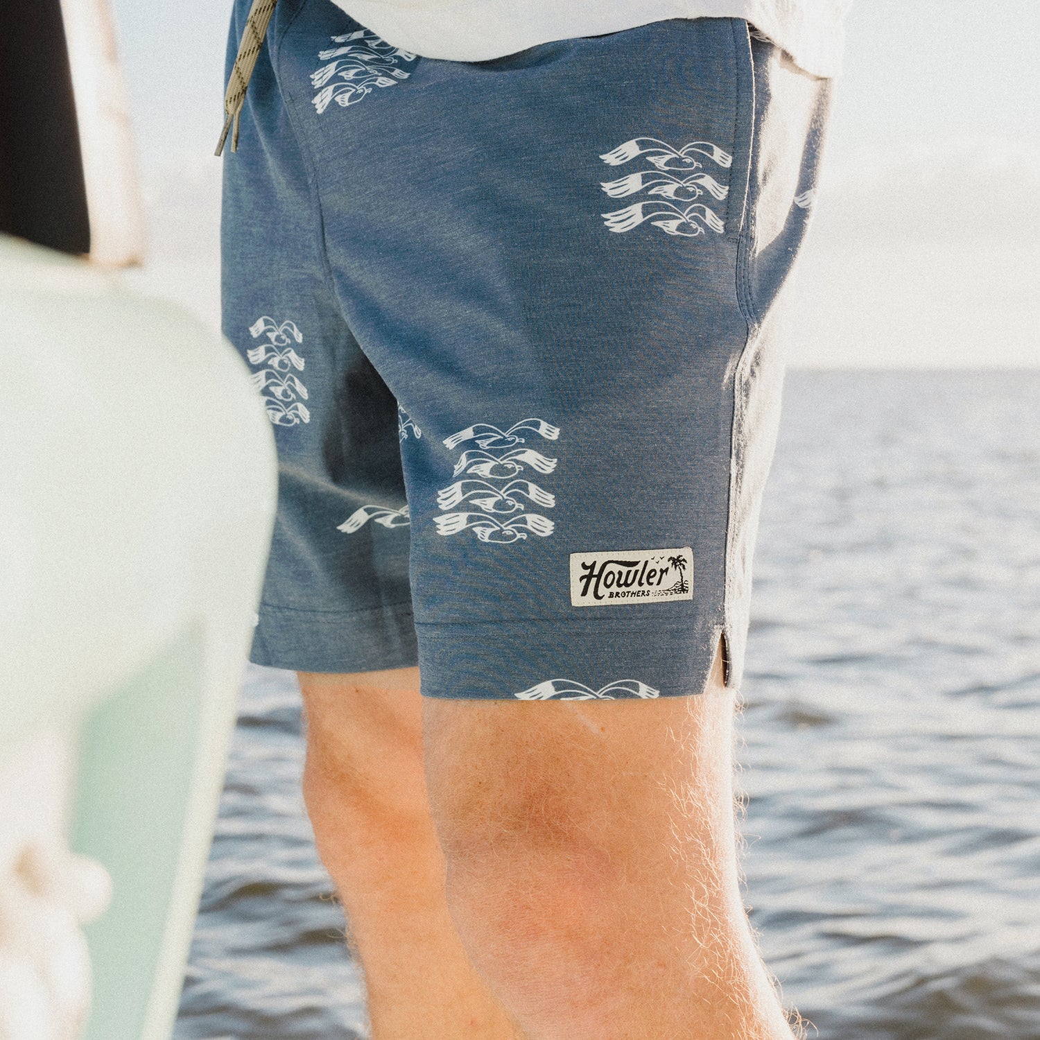 A person wearing blue Howler brand boardshorts with a 7” inseam and white wave patterns stands near the water on a sunny day.