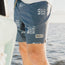 A person wearing blue Howler brand boardshorts with a 7” inseam and white wave patterns stands near the water on a sunny day.
