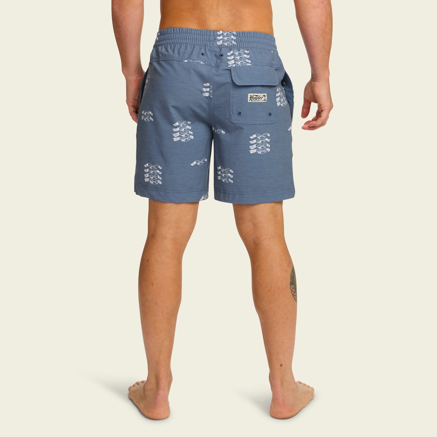 A person stands barefoot facing away, wearing blue boardshorts with a white wave pattern, a back pocket, and an elastic waistband. Their legs are visible with a small tattoo on the left calf. The 7” inseam shorts pop against the plain off-white background. - View 4