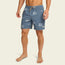 A man wearing blue boardshorts with a white wave pattern and 7” inseam stands barefoot against a plain light background. The boardshorts feature an elastic waistband with a drawstring and a small logo patch on the left leg.