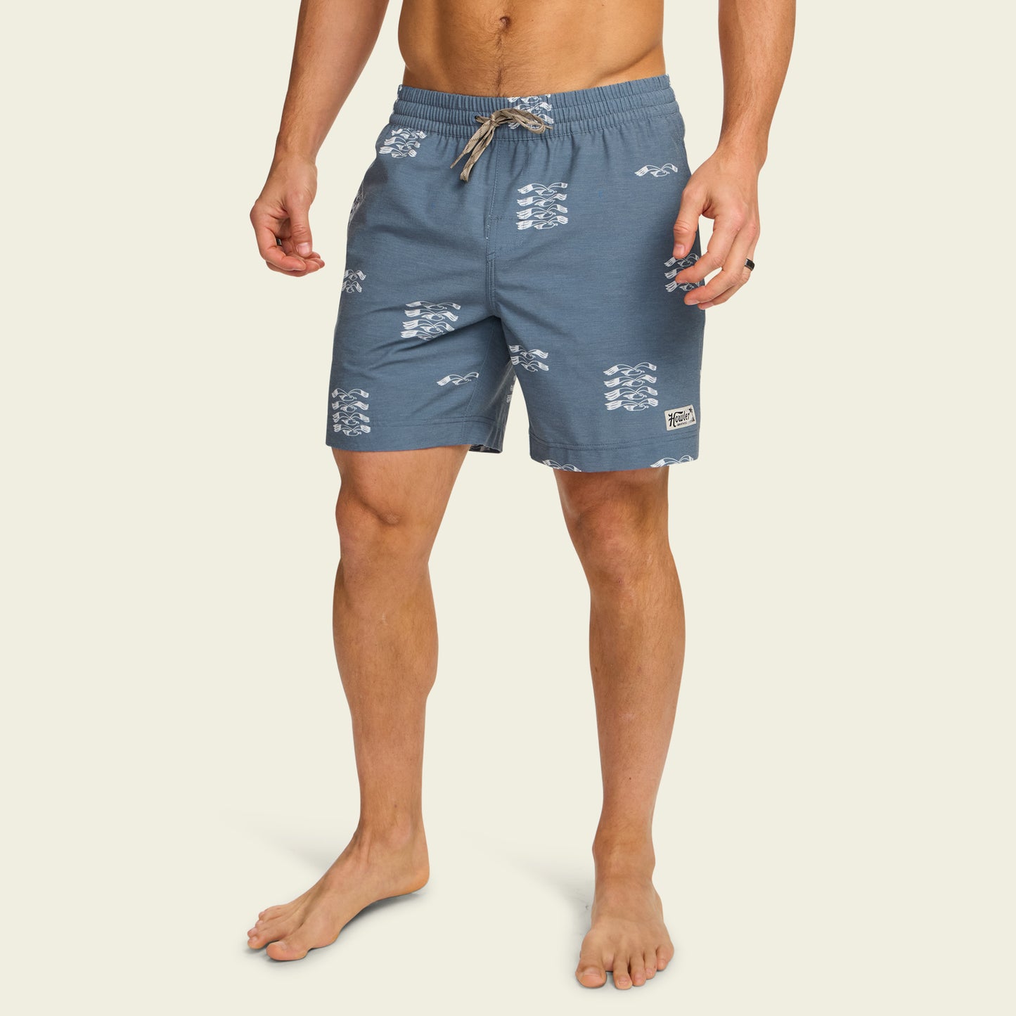 A man wearing blue boardshorts with a white wave pattern and 7” inseam stands barefoot against a plain light background. The boardshorts feature an elastic waistband with a drawstring and a small logo patch on the left leg. - View 3