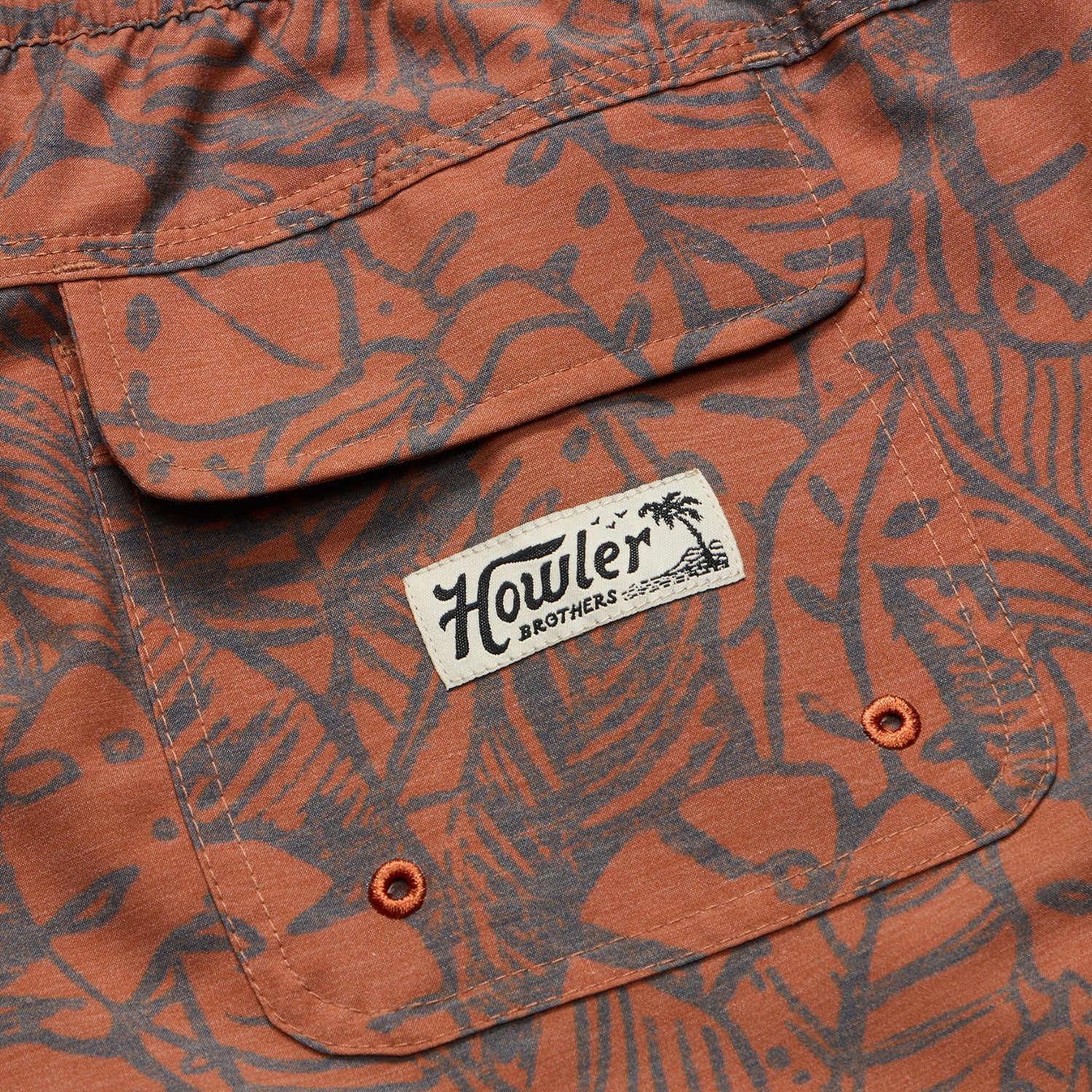 Close-up of rust-orange and dark gray patterned boardshorts with a pocket. The pocket features a sewn white Howler Brothers label with a palm tree graphic, two small orange eyelets at the bottom, and an elastic waistband for comfort. - View 5