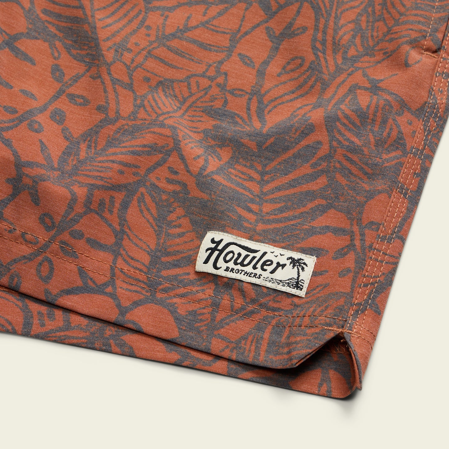 Close-up of a rust-orange fabric with a dark leaf and plant pattern, featuring a stitched Howler Brothers brand tag on the hem—perfect for boardshorts with an elastic waistband. - View 6