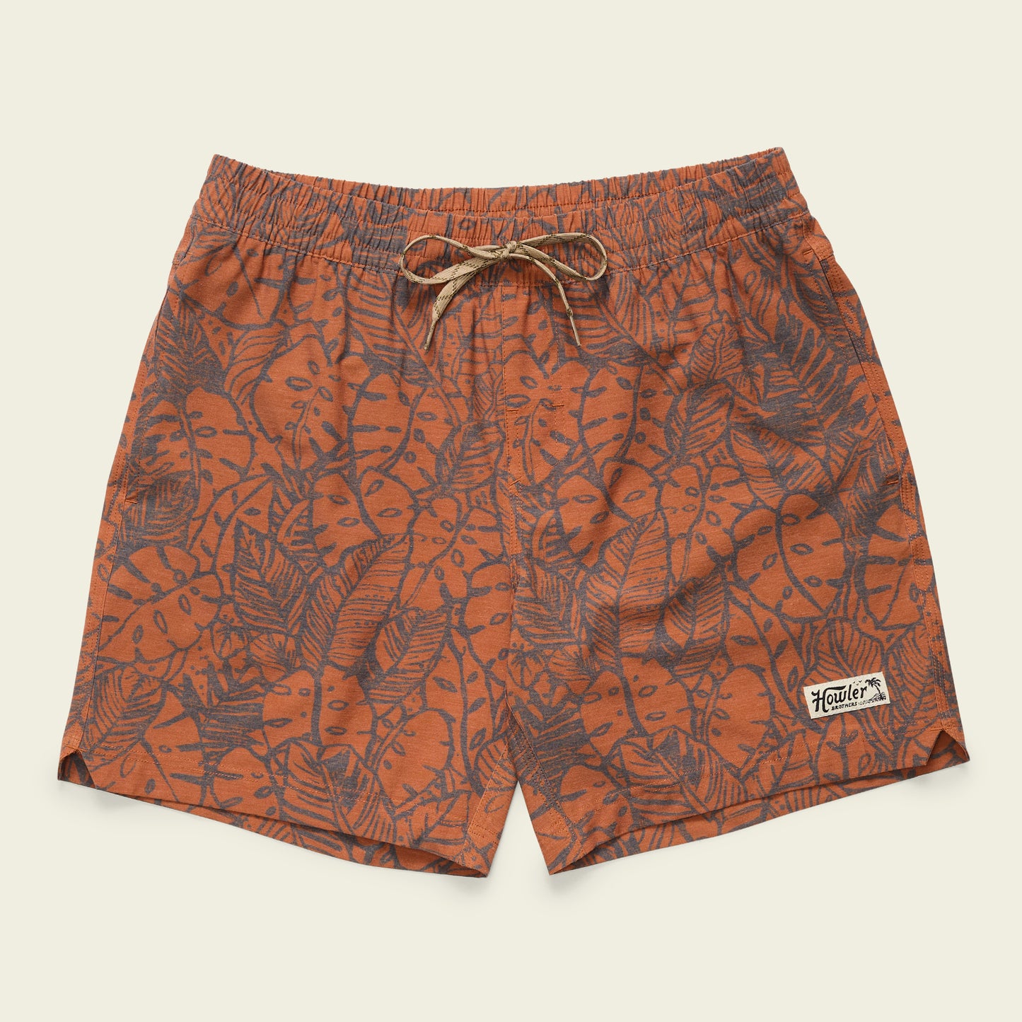 Orange swim shorts with a dark tropical leaf pattern, featuring a 7” inseam, elastic waistband with beige drawstring, and a small rectangular “Howler” patch sewn near the hem on the left leg. - View 1