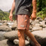A person stands on large rocks by a river, wearing a dark t-shirt and patterned peach boardshorts with a Howler patch and elastic waistband, surrounded by lush green trees.