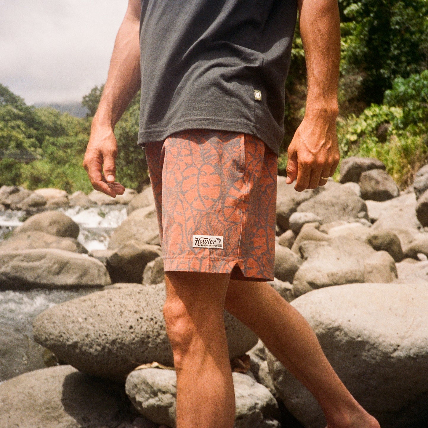 A person stands on large rocks by a river, wearing a dark t-shirt and patterned peach boardshorts with a Howler patch and elastic waistband, surrounded by lush green trees. - View 2