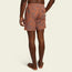 A person stands barefoot facing away, wearing orange boardshorts with a leafy pattern, an elastic waistband, and a 7” inseam. The shorts feature a small back pocket with a visible label.