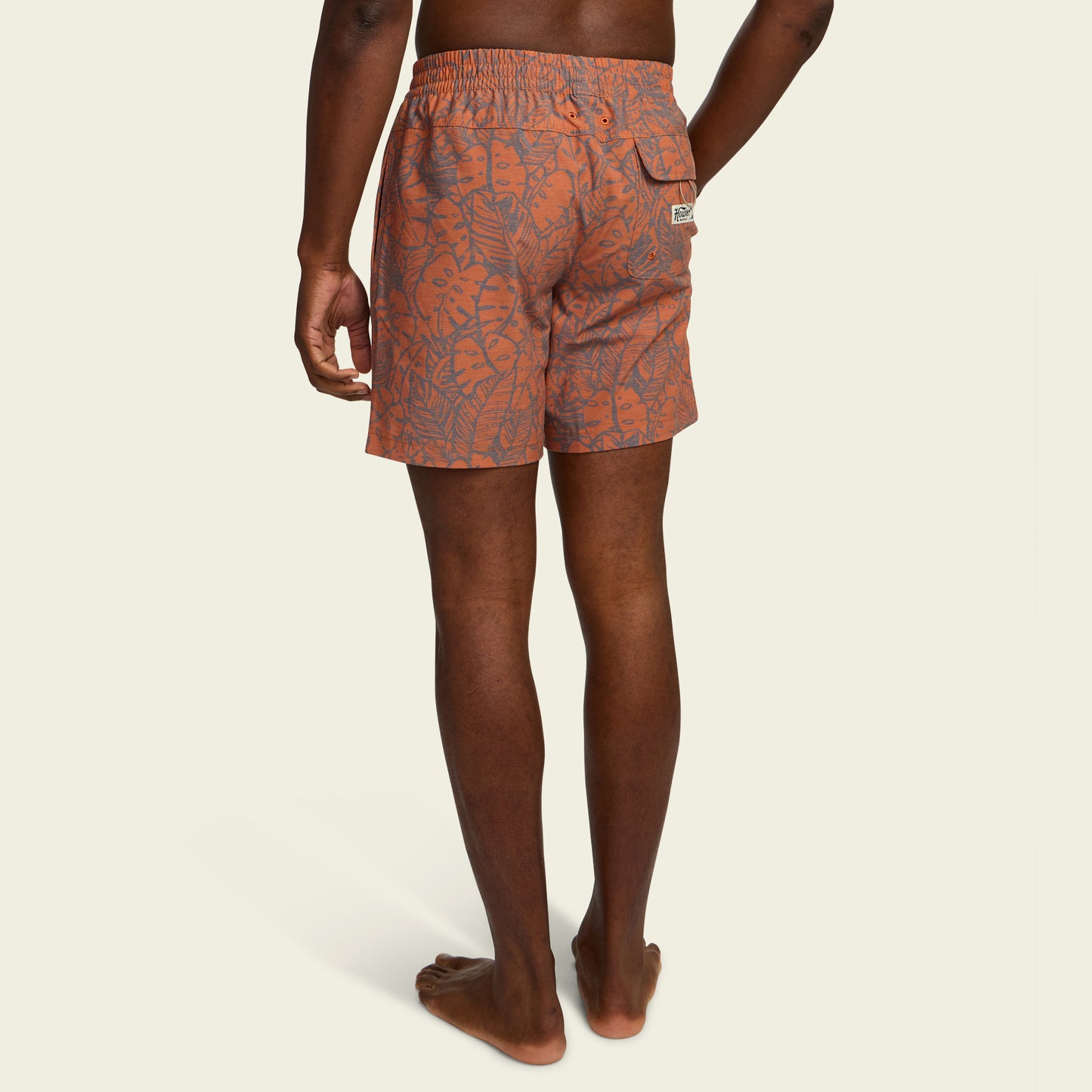A person stands barefoot facing away, wearing orange boardshorts with a leafy pattern, an elastic waistband, and a 7” inseam. The shorts feature a small back pocket with a visible label. - View 4