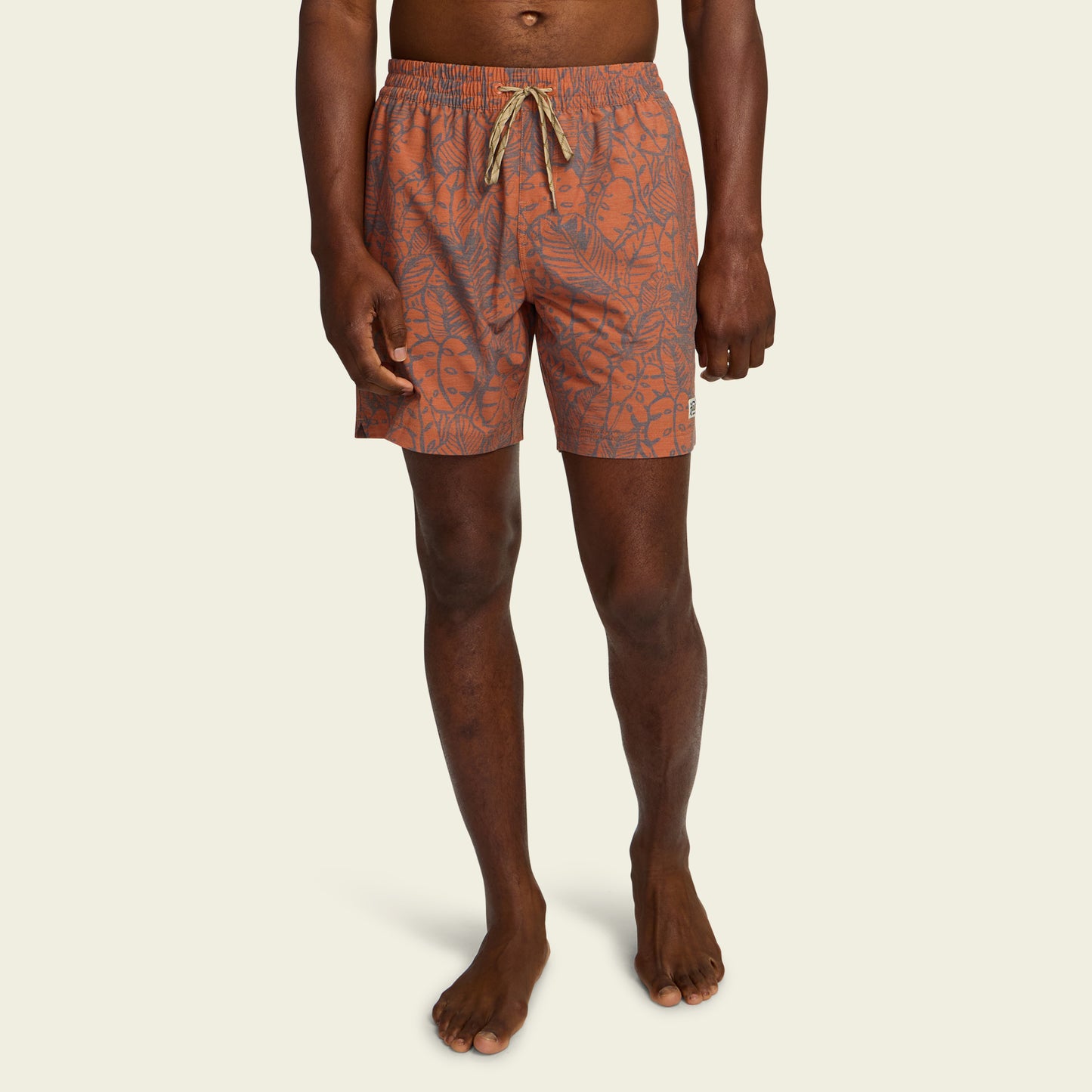A person wearing orange boardshorts with an abstract leaf pattern, a 7” inseam, and a drawstring waistband, standing barefoot against a plain light background. - View 3