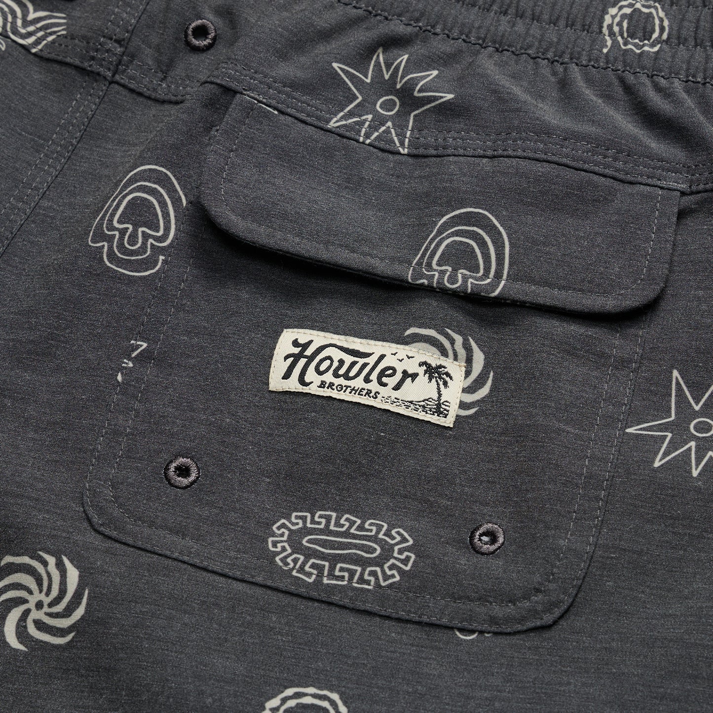 Close-up of a dark gray boardshorts pocket with abstract white patterns and a stitched Howler Brothers label under a palm tree graphic. The pocket, part of 7” inseam shorts, features two button holes at the bottom. - View 5