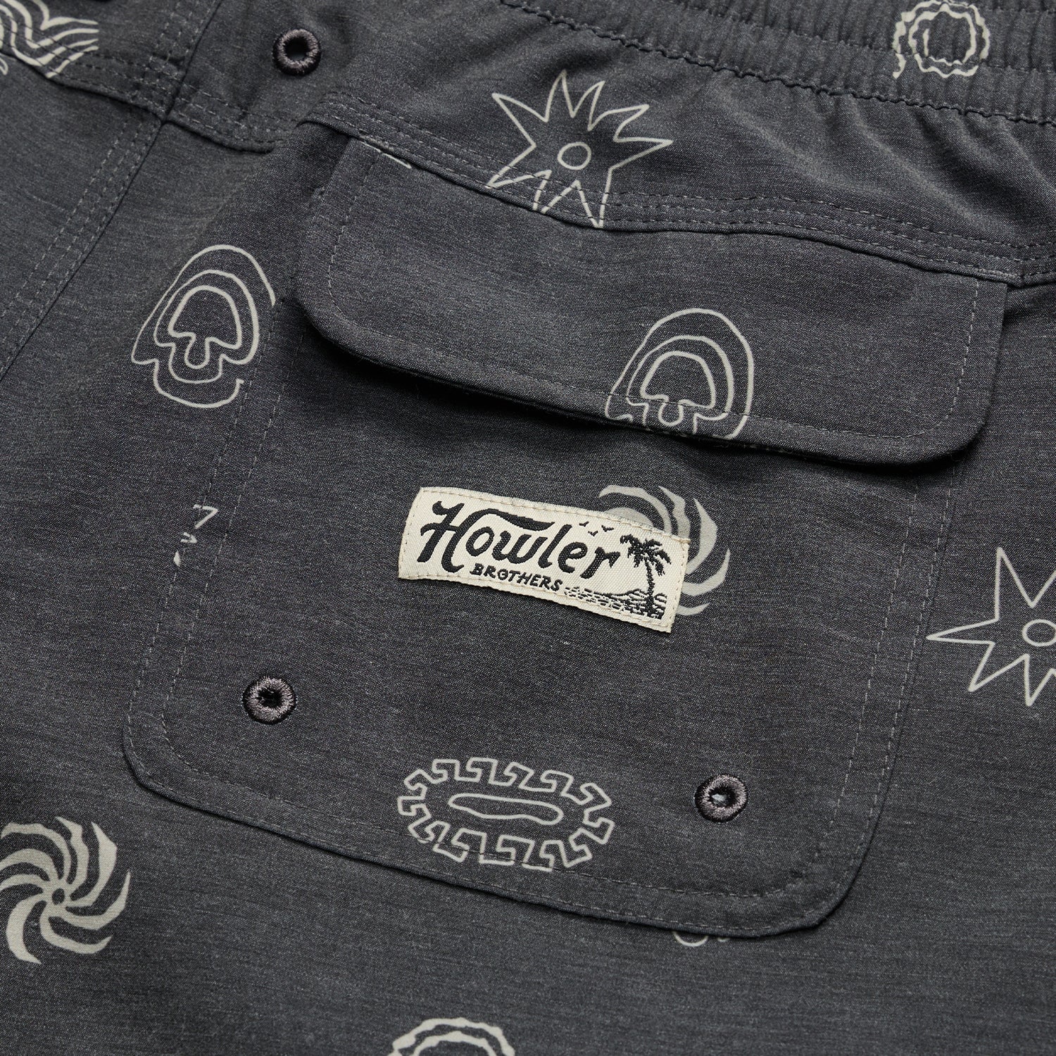 Close-up of a dark gray boardshorts pocket with abstract white patterns and a stitched Howler Brothers label under a palm tree graphic. The pocket, part of 7” inseam shorts, features two button holes at the bottom. - View 5