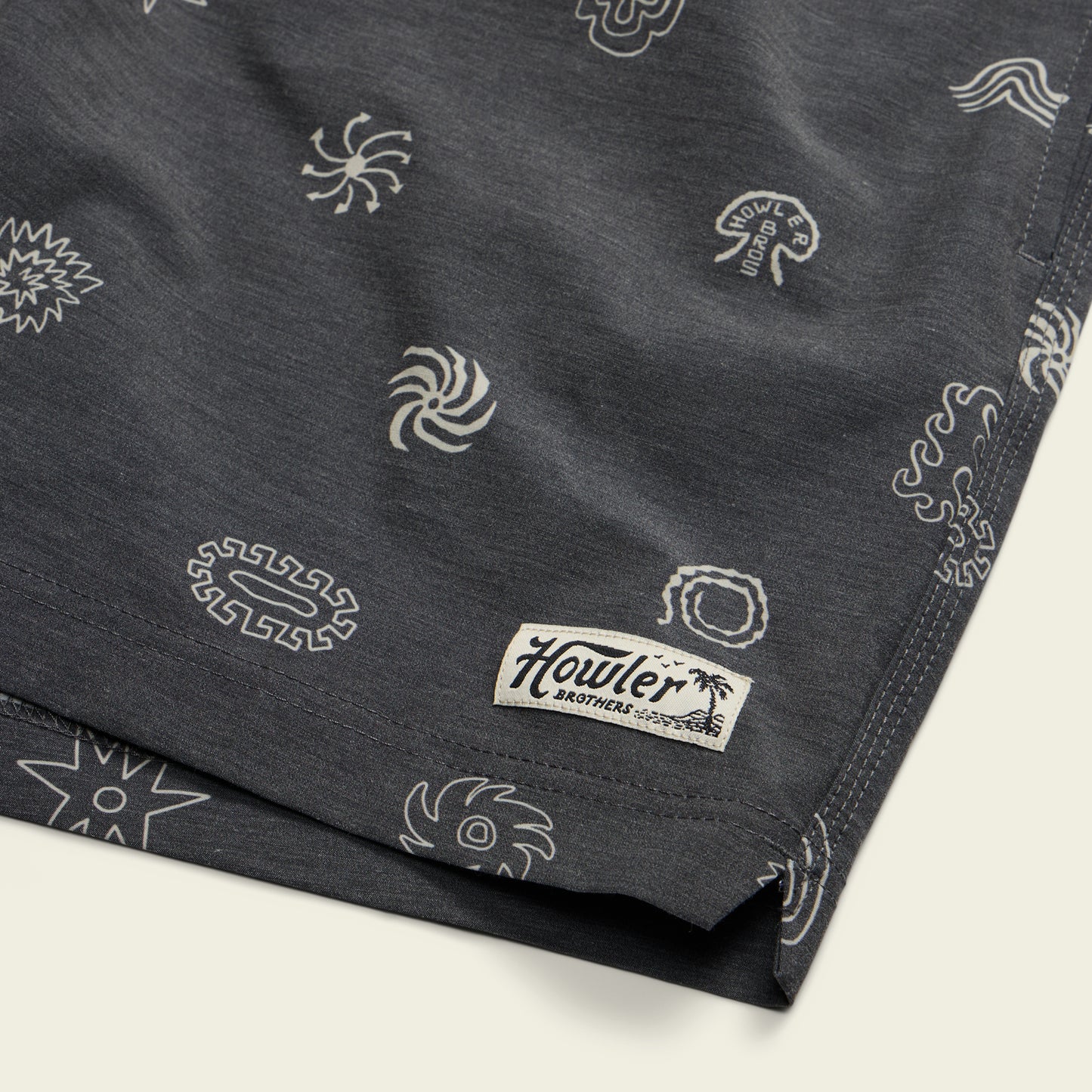 Dark boardshorts with white abstract and sun-like designs, featuring a 7” inseam and an elastic waistband. Finished with a small rectangular Howler Brothers label with a palm tree near the hem. - View 6