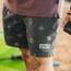 A person stands outdoors wearing boardshorts with a 7” inseam, featuring white sun and wave designs on dark fabric. Part of a purple shirt is visible, and they are holding a black object in their right hand.