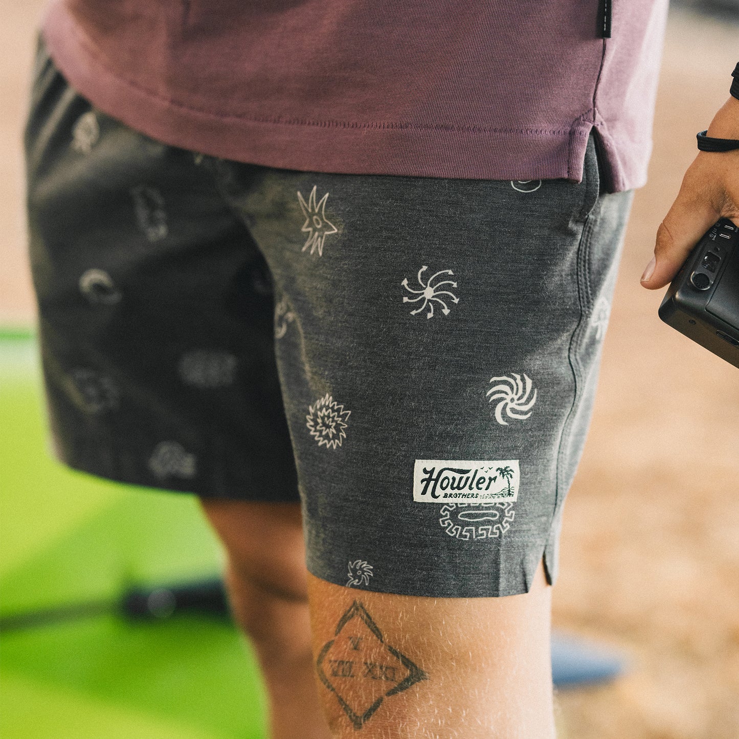 A person stands outdoors wearing boardshorts with a 7” inseam, featuring white sun and wave designs on dark fabric. Part of a purple shirt is visible, and they are holding a black object in their right hand. - View 2