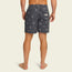 A man stands barefoot facing away, wearing dark gray boardshorts with a subtle pattern, a 7” inseam, an elastic waistband, and a back pocket with a white label, against a plain light background.