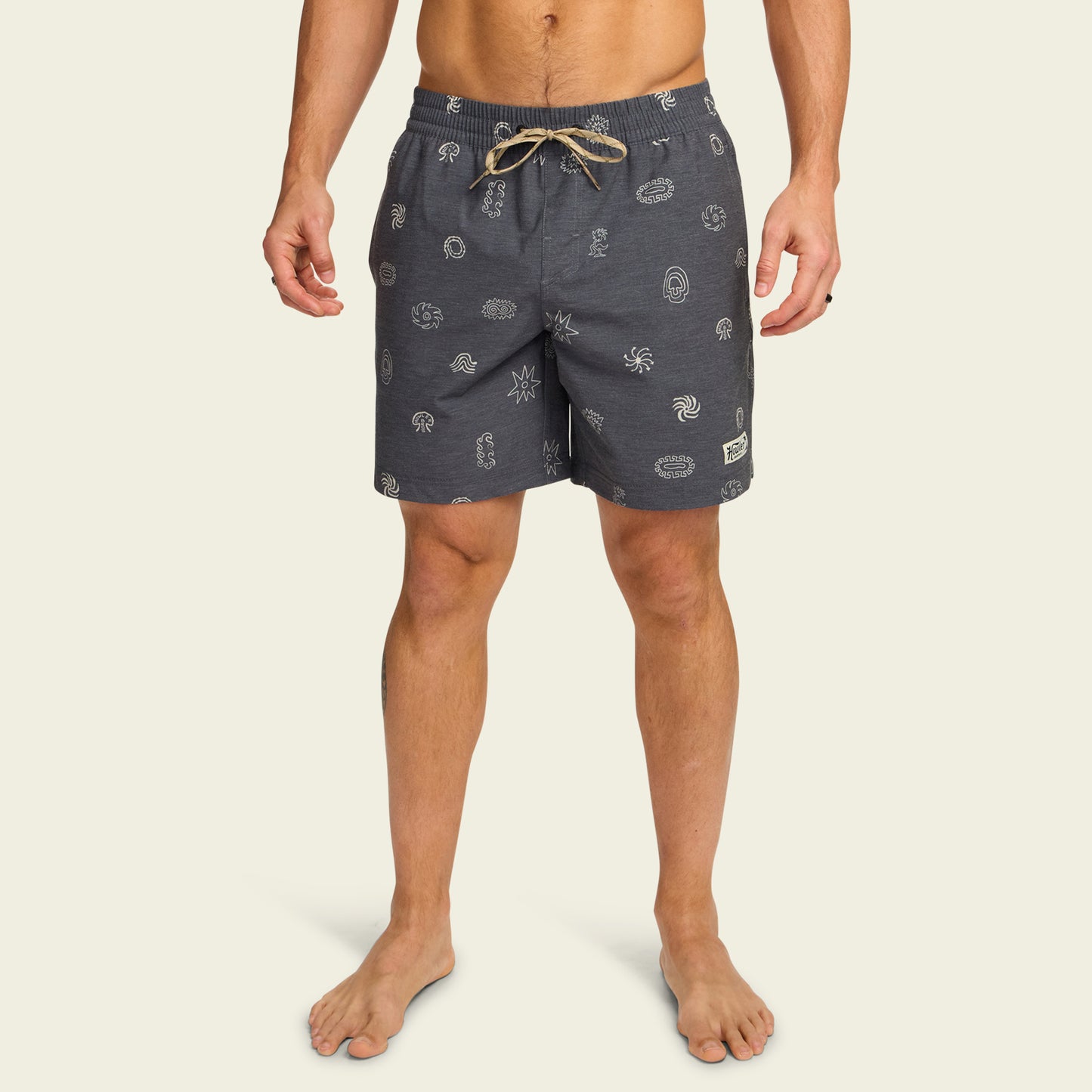 A man wearing navy blue boardshorts with white printed patterns and a beige drawstring stands barefoot against a plain light background. These boardshorts feature an elastic waistband and a comfortable 7” inseam. - View 3
