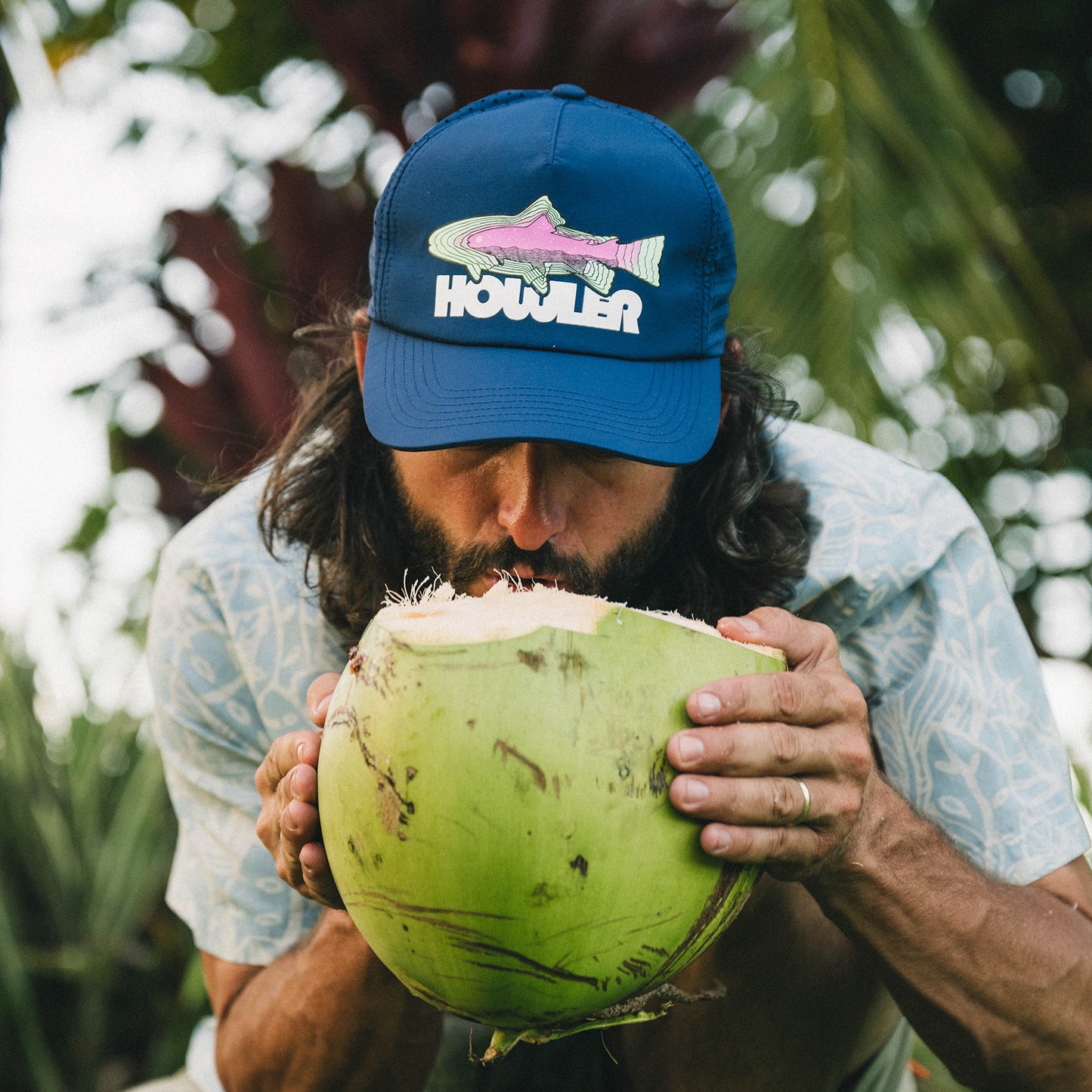 A man with long hair and a beard drinks from a green coconut while wearing a blue breathable tech hat with a fish graphic and the word “Howler.” He is outdoors, surrounded by lush tropical plants, ready for outdoor adventures. - View 2