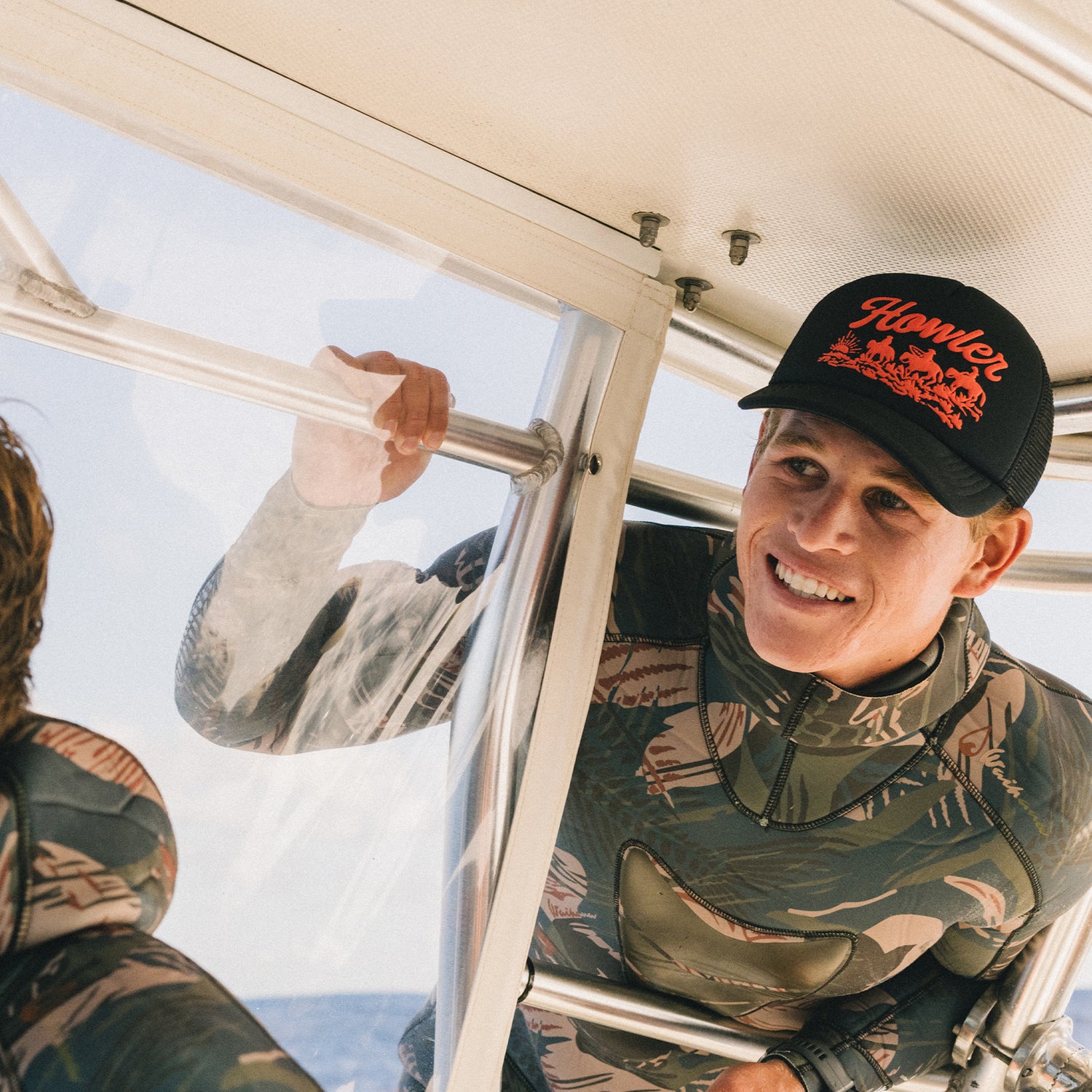 A smiling person in camouflage clothing and a black vintage-inspired hat holds onto a metal bar inside a boat, with sunlight streaming in and another person partially visible beside them.