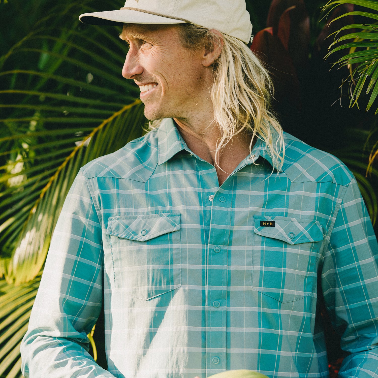 A smiling man with long blonde hair wearing a lightweight stretch shirt and a beige cap stands outdoors in front of lush green palm leaves, looking to the side.