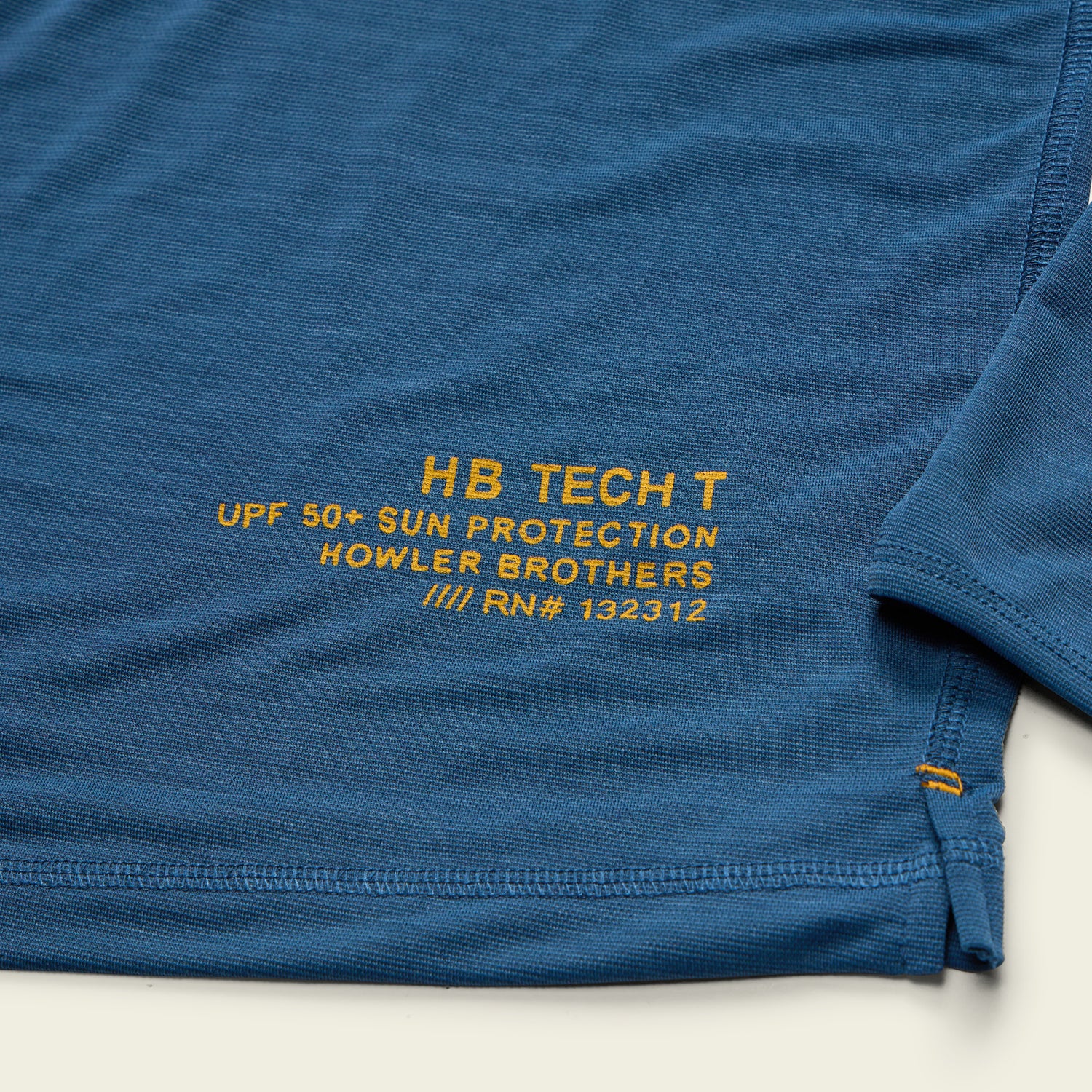 A close-up of a blue hooded tech shirt with yellow text reading HB TECH T UPF 50+ SUN PROTECTION HOWLER BROTHERS /// RN# 132312 near the bottom hem. Visible stitching detail and part of the sleeve. - View 6