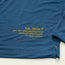 A close-up of a blue hooded tech shirt with yellow text reading HB TECH T UPF 50+ SUN PROTECTION HOWLER BROTHERS /// RN# 132312 near the bottom hem. Visible stitching detail and part of the sleeve.