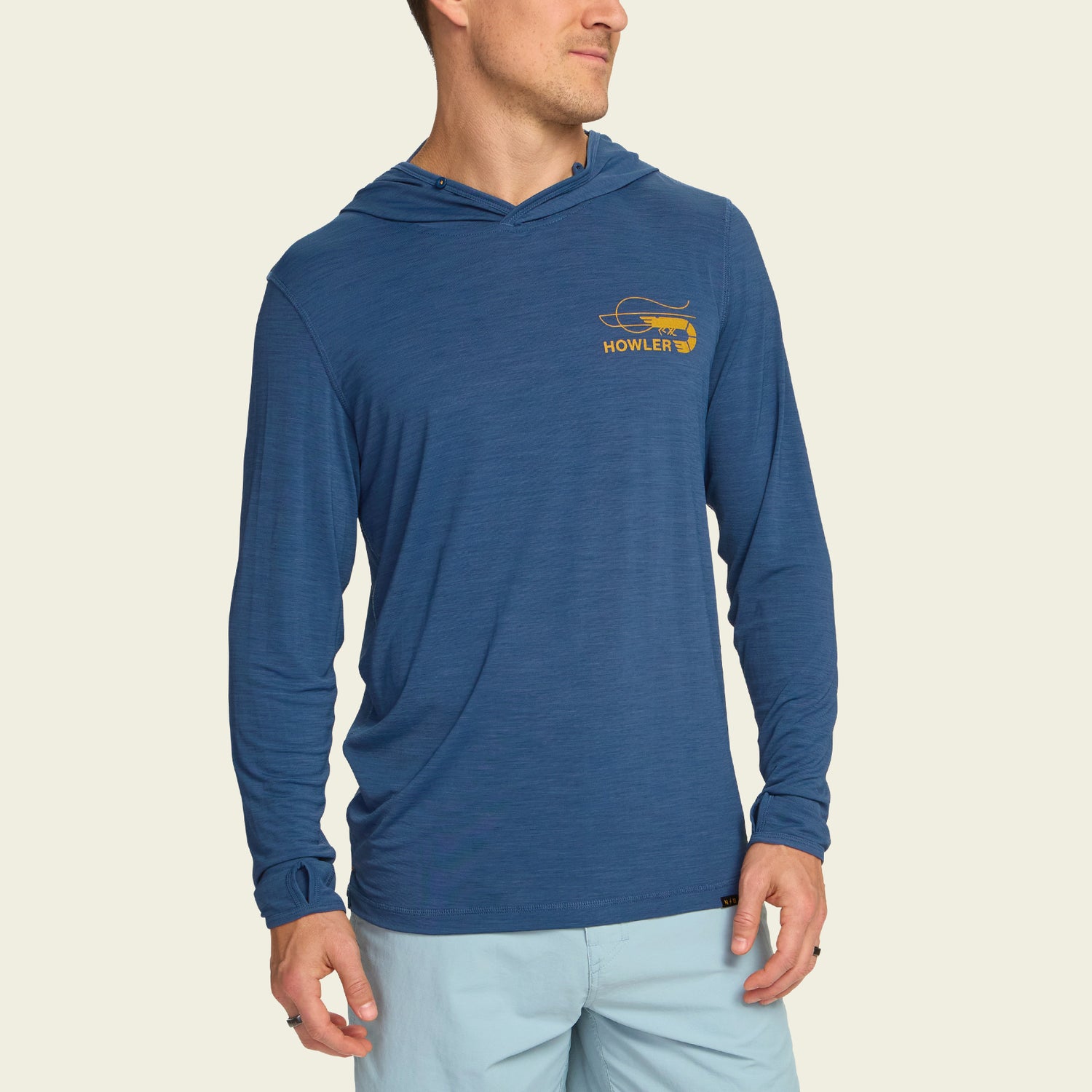 A man wearing a blue lightweight hoodie with a small yellow Howler logo and graphic on the left chest, paired with light blue shorts, stands against a plain off-white background. - View 4