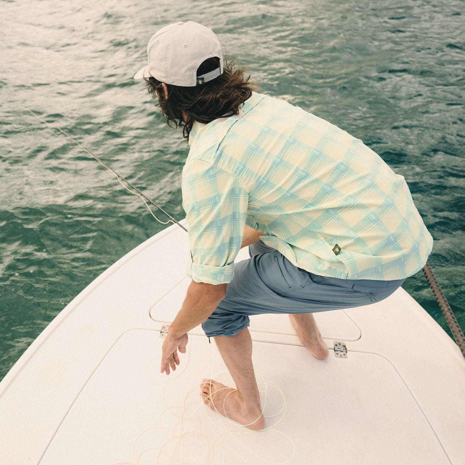 A person in a light Matagorda Shirt, blue pants, and a light cap stands barefoot on a boat, holding fishing line over turquoise water, facing away from the camera.