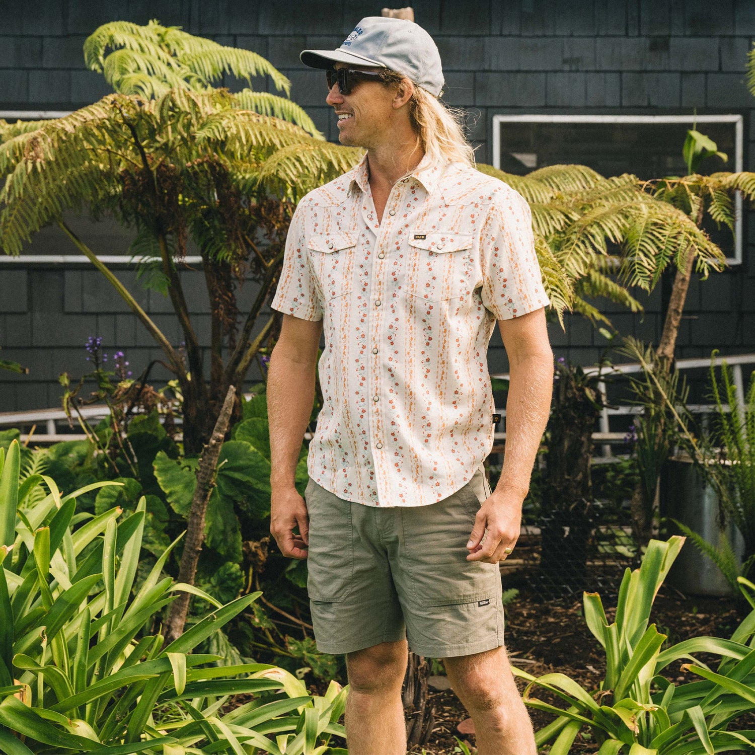 A man wearing a light patterned short-sleeve shirt, Quonset Camp Shorts with vintage-style buttons, and a gray cap stands smiling among green plants in front of a dark building with large windows.