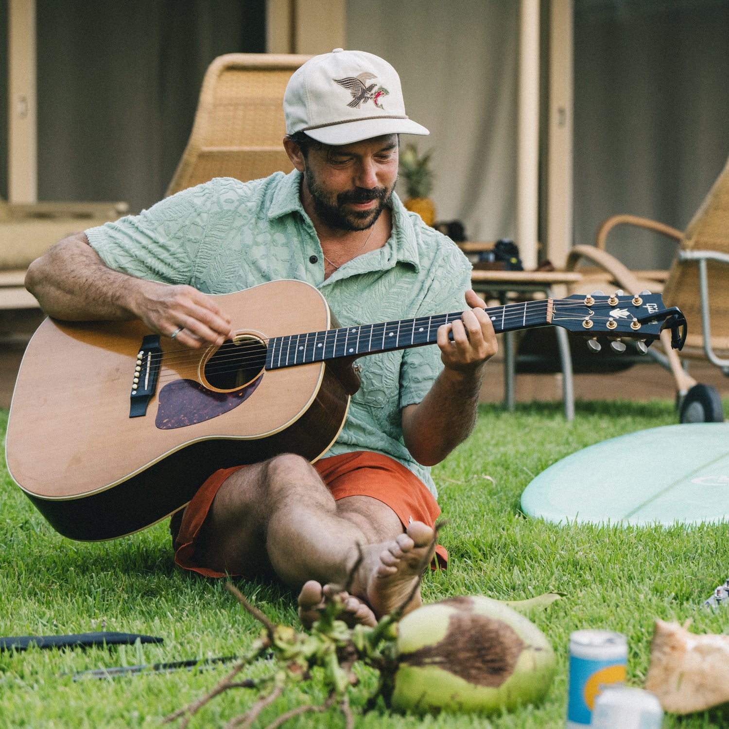 A man sitting barefoot on grass plays an acoustic guitar, wearing a light green terry cloth polo, orange shorts, and a white cap. Nearby are a surfboard, coconut, branches, drinks, wicker chairs, and a building in the background.