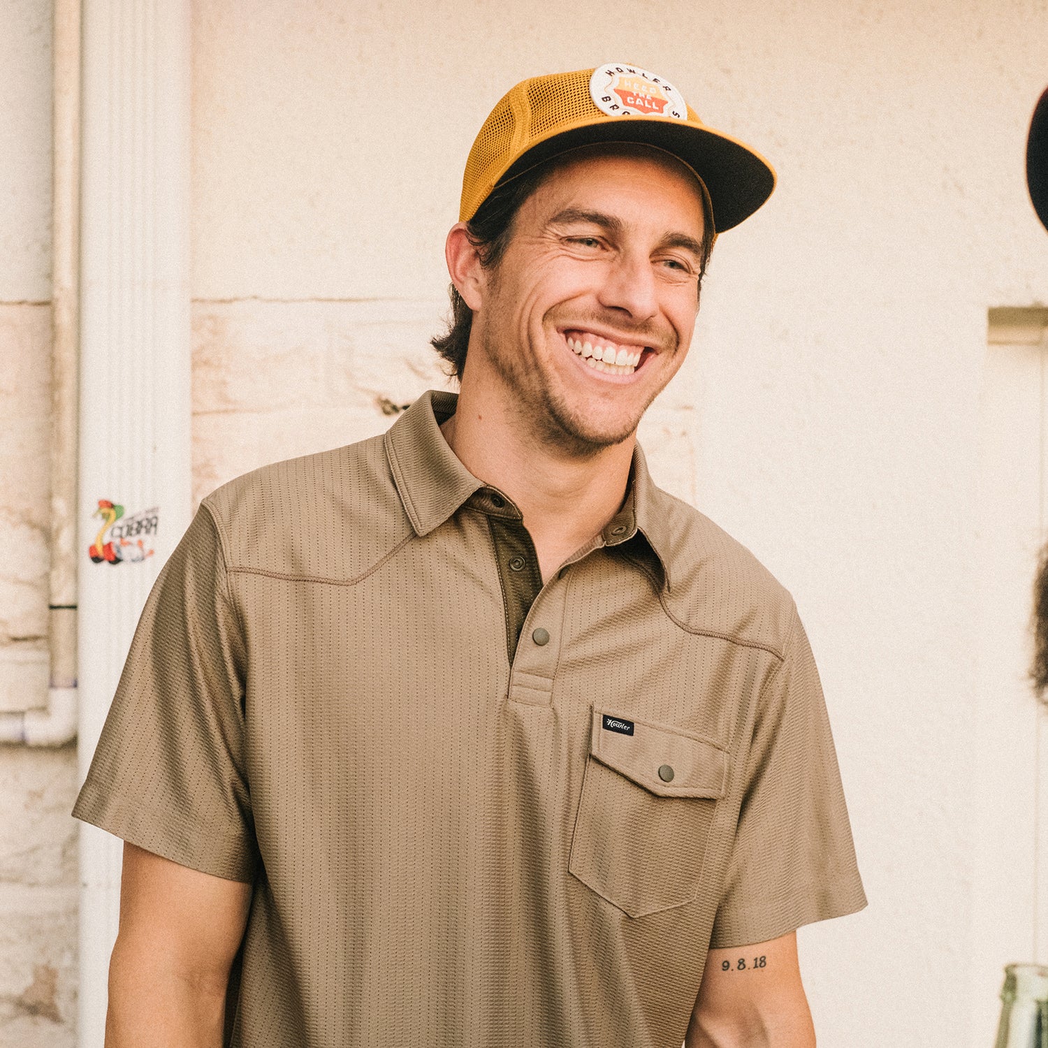 A smiling man in a tan tech polo and mustard yellow cap stands outdoors. He has a tattoo with numbers on his right forearm and is positioned in front of a light-colored wall with pipes and a sticker.