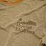 A close-up of a tan polyester spandex blend shirt with the Howler Brothers logo printed in dark brown, featuring stylized text below a minimalist landscape graphic with a sun and mountains.