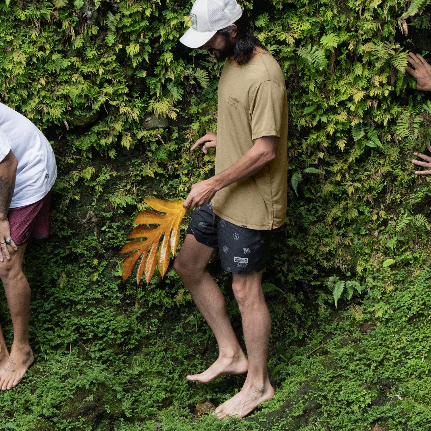 A barefoot person in a lightweight UPF shirt and black shorts holds a large yellow leaf, standing on lush green ground near a wall covered in ferns. Other people stand nearby, their bodies partially out of frame.