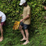 A barefoot person in a lightweight UPF shirt and black shorts holds a large yellow leaf, standing on lush green ground near a wall covered in ferns. Other people stand nearby, their bodies partially out of frame.