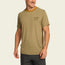 A smiling man wears a light brown Howler Brothers shortsleeve tech shirt with a mustard-colored neckline and dark shorts, standing against a plain off-white background.