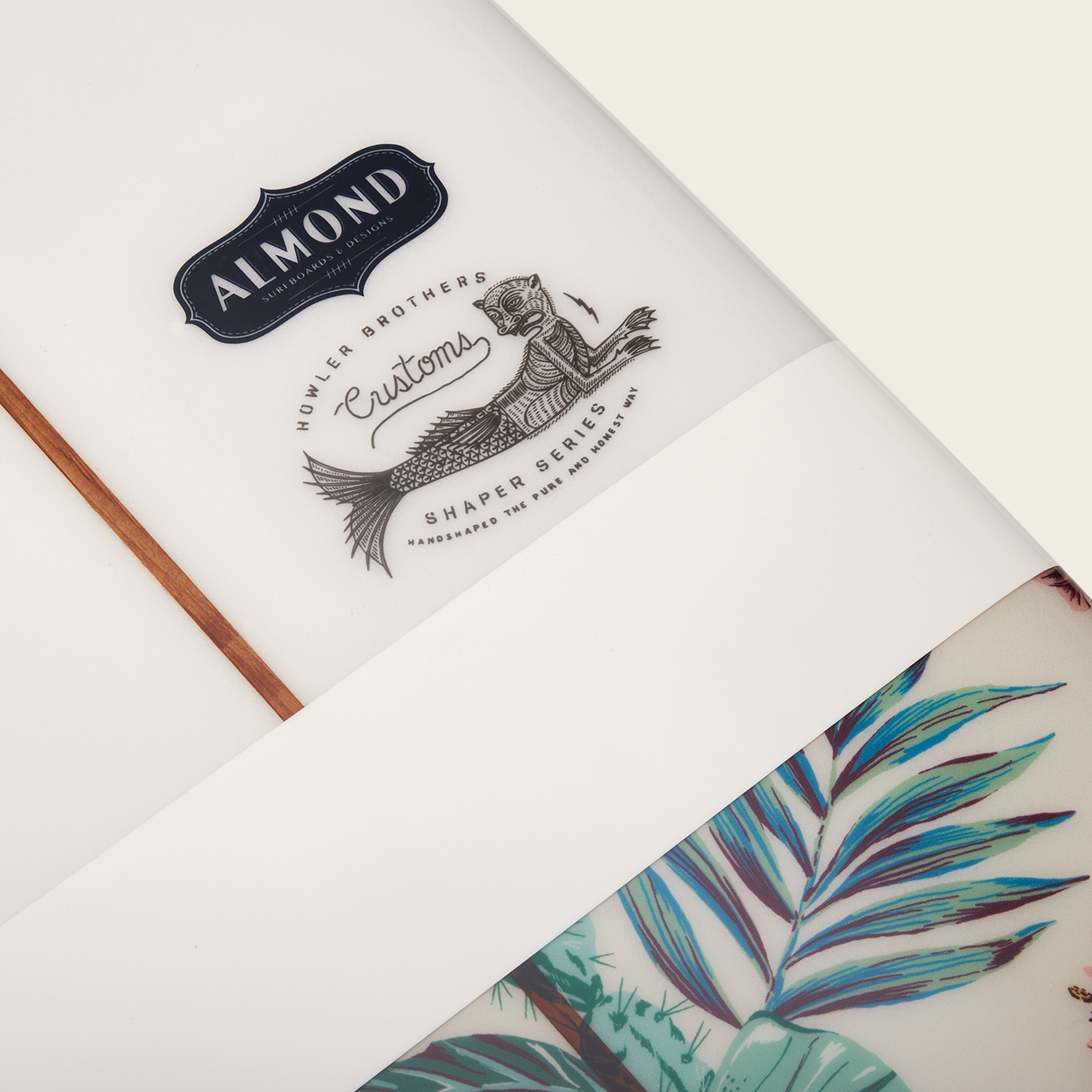 Close-up of a Surf Thump longboard featuring the Almond brand logo, a mermaid illustration labeled “Hokler Brothers Customs Shaper Series,” and part of a tropical leaf design, highlighting its versatile handling.